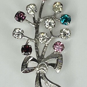 May include: Silver-toned brooch in a floral design with a bow. The brooch features a branch-like structure with colorful, round gemstones in red, purple, pink, teal, and clear. A decorative accessory for clothing.