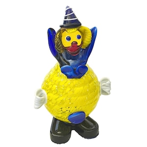 May include: A whimsical glass clown figurine. The clown has a yellow body with a textured pattern, blue arms, and a black hat. The face is yellow with blue eyes and a red mouth. White accents are on the sides.
