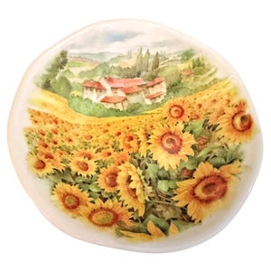 May include: An off-white ceramic plate with a painted landscape of sunflowers and a rural Italian village. The sunflowers are yellow and orange, with green leaves. The village has red-tiled roofs. The plate is round.