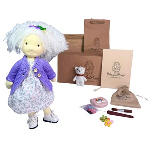 May include: A fabric doll with white yarn hair, wearing a purple knit cardigan over a floral dress and white shoes. Included are a small white bear, a brown paper bag, a card, a drawstring pouch, a rainbow case, and accessories labeled BlissfulPixie.