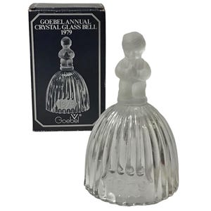 May include: A clear glass bell with a frosted figure on top and vertical ribbed detailing. The bell is next to a black box with the text "GOEBEL ANNUAL CRYSTAL GLASS BELL 1979". The bell has the year 1979 etched on it.