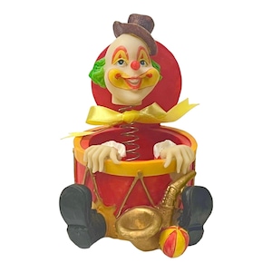 Puede incluir: Un juguete Jack-in-the-box caprichoso, rojo y amarillo, con un payaso sonriente. El payaso tiene pelo verde, un sombrero marrón y un lazo amarillo. Sostiene un saxofón y una pelota roja y amarilla. Un artículo decorativo divertido.