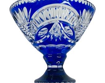 May include: A cobalt blue crystal bowl with a pedestal base. The bowl features intricate cut glass patterns and a scalloped rim. The bowl is approximately 8 inches in diameter and 6 inches tall, perfect for display or serving.