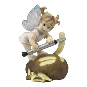 May include: A figurine of a fairy with blonde pigtails, blue accents, and translucent wings, holding a silver rolling pin over a potato. The fairy is wearing a white outfit and stands on a brown potato-shaped base.