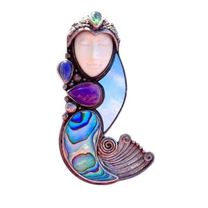 May include: A handcrafted silver pendant featuring a serene face carved from bone, adorned with a green gemstone, blue and purple stones, and iridescent abalone shell. The pendant has a unique, artistic design.