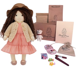 May include: A fabric doll with long brown hair, wearing a straw hat, a beige knit cardigan with two buttons, and a peach-colored dress. Included are a small white bear, a brown paper bag, a card, a drawstring pouch, and sewing accessories.