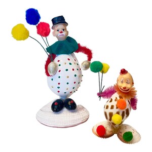 May include: Two clown figurines made from seashells. One is white with red and green polka dots, holding pom-pom balloons. The other is brown and white with orange and yellow pom-poms. Both have painted faces and wear hats.