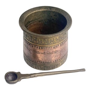 May include: An antique metal container and spoon. The cylindrical container features decorative bands and etched text. The spoon has a small, rounded bowl and a long handle. The metal has a tarnished, aged appearance, suggesting age.