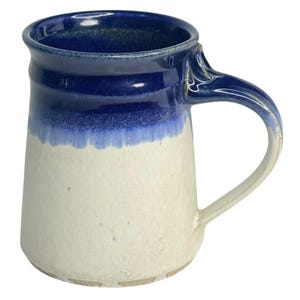 May include: A handmade ceramic mug with a white base and a blue gradient top. The mug has a curved handle and a slightly flared rim. The blue color transitions from a dark shade at the top to a lighter shade towards the middle.