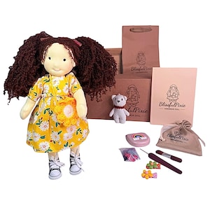 May include: A fabric doll with curly brown hair wearing a yellow floral dress and black and white sneakers, accompanied by a small white bear, a brown paper bag, a pink box, a beige pouch, a rainbow case, hair accessories, and a tool.