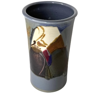 May include: A tall, ceramic vase with a wide opening and a cylindrical shape. The vase features a hand-painted design with abstract shapes in shades of brown, blue, and cream. The rim and base are a muted blue, and the overall aesthetic is rustic.