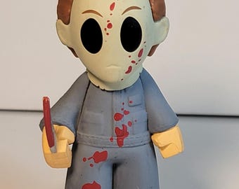 May include: A stylized figure of Michael Myers from the Halloween horror film series. The figure wears a blue jumpsuit and a white mask with black eyes and red blood splatters. It holds a red knife.