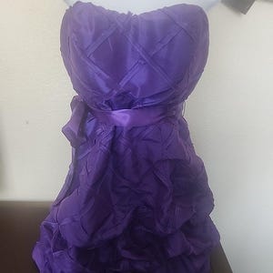 May include: A strapless, knee-length purple dress. The bodice features a quilted design, and a satin ribbon belt cinches the waist. The skirt has a layered, ruffled effect. The dress is displayed on a white mannequin bust.