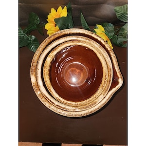 May include: A set of three nested ceramic mixing bowls featuring a glossy dark brown interior and a textured, cream-colored drip glaze along the outer rims. The bowls are stacked vertically, showcasing a consistent circular design and smooth finish.