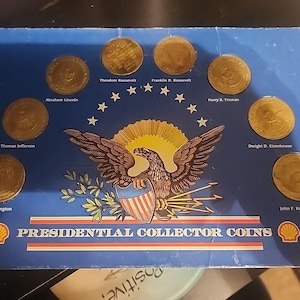 Può includere: Un espositore blu con otto monete da collezione presidenziali color oro. Le monete sono disposte attorno a un emblema centrale di un'aquila con uno scudo. I nomi dei presidenti sono stampati sotto ogni moneta.