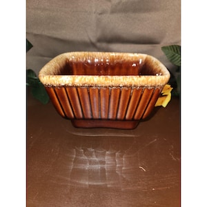 May include: A square ceramic bowl featuring a deep brown glaze with vertical ribbed detailing along the exterior sides. The rim is finished with a lighter, cream-colored drip glaze effect that contrasts with the darker body of the vessel.