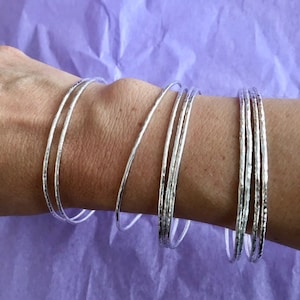 Peut inclure: Un ensemble de bracelets en argent avec une texture martelée. Les bracelets sont empilés sur le poignet d'une personne.