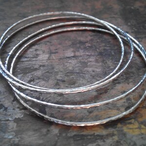 May include: Four silver bangles stacked on top of each other. The bangles are hammered and have a textured surface.