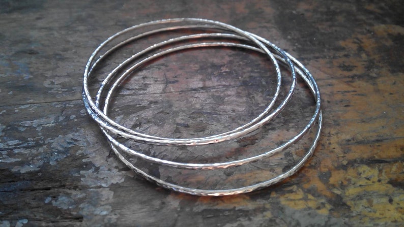 May include: Four silver bangles stacked on top of each other. The bangles are hammered and have a textured surface.