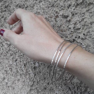 May include: Five silver bangles stacked on a wrist. The bangles are thin and have a hammered texture.