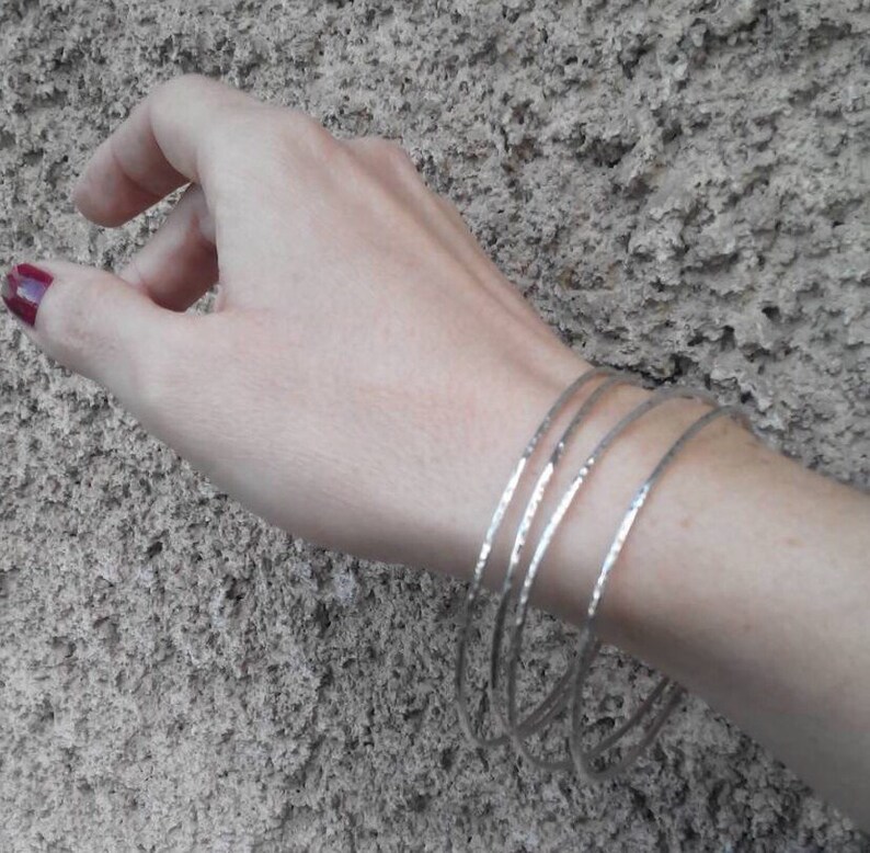 May include: Five silver bangles stacked on a wrist. The bangles are thin and have a hammered texture.