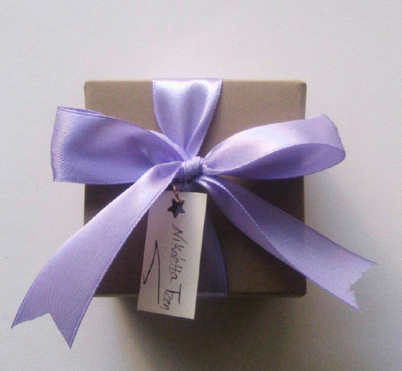 May include: A small brown gift box with a lavender satin ribbon tied in a bow. A white tag with the text "Mikelle Tzen" is attached to the ribbon.