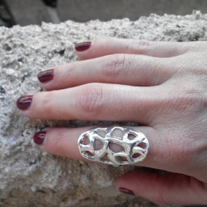 May include: A silver ring with an open, web-like pattern. The ring is oval and worn on a finger. The hand has dark red nail polish. The background is a rough, textured surface, possibly stone or concrete.