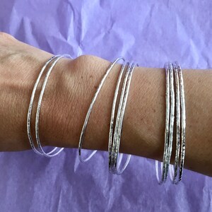 May include: A set of silver bangles with a hammered texture. The bangles are stacked on a person's wrist.