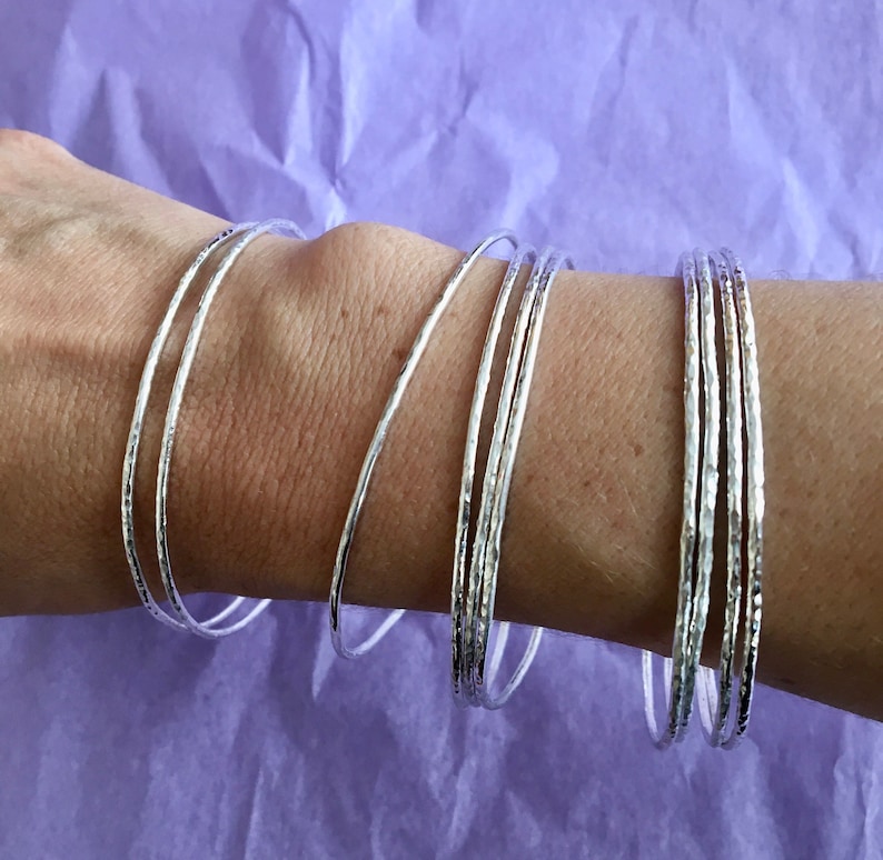 May include: A set of silver bangles with a hammered texture. The bangles are stacked on a person's wrist.