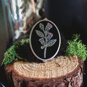 May include: An oval-shaped, silver-toned pendant with a black background, featuring a detailed floral design. The flower has purple and green accents. The pendant is set against a backdrop of moss and a wood slice.