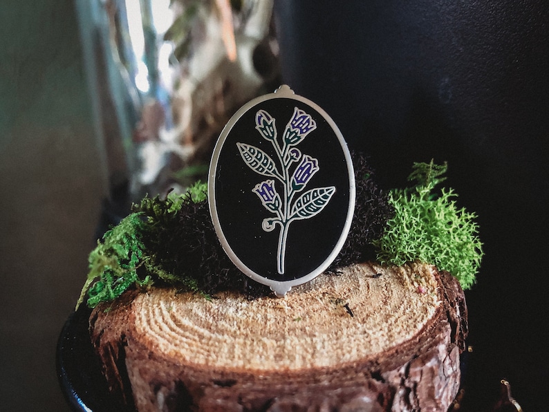 May include: An oval-shaped, silver-toned pendant with a black background, featuring a detailed floral design. The flower has purple and green accents. The pendant is set against a backdrop of moss and a wood slice.