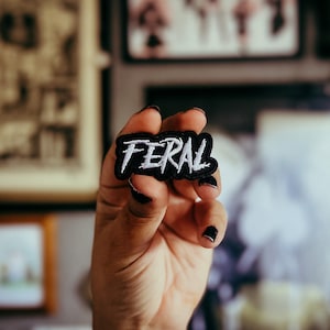 Puede incluir: Un parche negro con la palabra "FERAL" bordada en blanco, sostenido frente a un fondo borroso. El parche tiene un borde blanco y es de forma rectangular. La palabra está en una fuente audaz y estilizada.