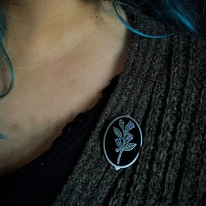 May include: An oval-shaped pin with a black background and a silver border. The pin features a botanical design with green leaves and purple accents. The pin is attached to a dark gray sweater.