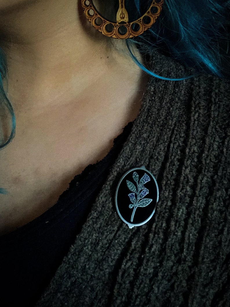 May include: An oval-shaped pin with a black background and a silver border. The pin features a botanical design with green leaves and purple accents. The pin is attached to a dark gray sweater.