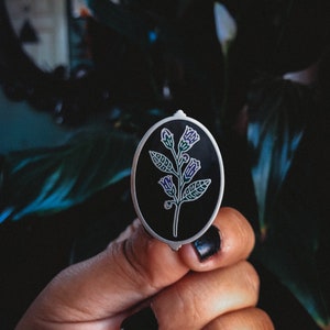 May include: An oval-shaped, silver-edged enamel pin featuring a stylized floral design. The pin has a black background with a white and purple floral illustration. The pin is held in a hand with black nail polish.