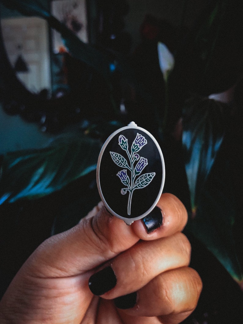 May include: An oval-shaped, silver-edged enamel pin featuring a stylized floral design. The pin has a black background with a white and purple floral illustration. The pin is held in a hand with black nail polish.