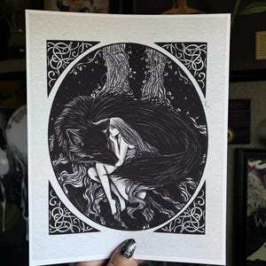 May include: Black and white art print featuring a woman embracing a wolf within a circular frame. The illustration depicts a woman with long hair, nestled against a wolf, surrounded by trees and stars. The border has intricate Celtic knot designs.