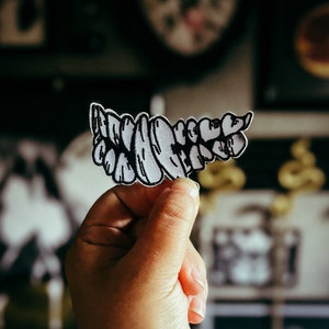 Puede incluir: Un parche en blanco y negro con forma de dientes. El parche está sostenido por una mano. Los dientes son blancos con contornos negros. El fondo está borroso, con varias imágenes enmarcadas visibles. Un accesorio único.