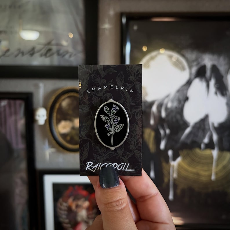 May include: An oval-shaped enamel pin featuring a botanical illustration of a flower with multiple blossoms and leaves in white and purple outlines against a solid black background, mounted on a black display card labeled ENAMELPIN RAICODOLL.