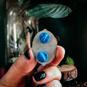 May include: Close-up of the back of an oval-shaped enamel pin with two blue pin backs. The pin has a silver-colored base with a textured surface. The pin is being held by a person with black nail polish.