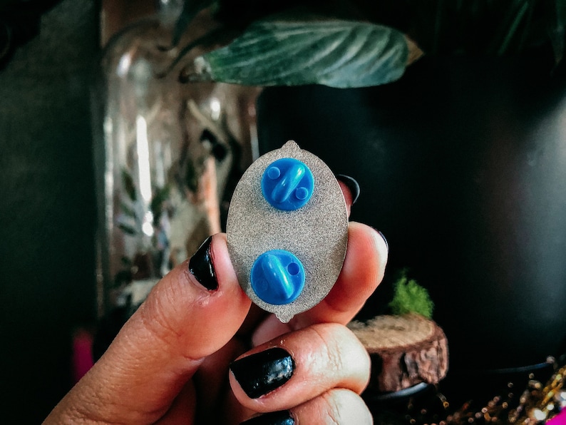 May include: Close-up of the back of an oval-shaped enamel pin with two blue pin backs. The pin has a silver-colored base with a textured surface. The pin is being held by a person with black nail polish.