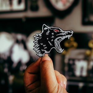 Puede incluir: Un parche bordado negro con la cabeza de un lobo con la boca abierta y dientes visibles. El lobo tiene ojos rojos y detalles blancos. El parche se sostiene contra un fondo borroso.