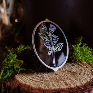May include: An oval pendant with a floral design. The pendant has a black background with a silver border. The floral design features blue and white flowers and green leaves. The pendant is resting on a wooden surface with green moss.
