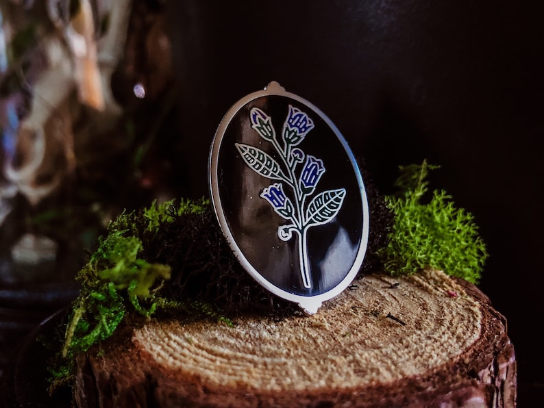 May include: An oval pendant with a floral design. The pendant has a black background with a silver border. The floral design features blue and white flowers and green leaves. The pendant is resting on a wooden surface with green moss.