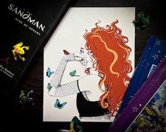 May include: A drawing of a person with red hair and butterflies, wearing a black top and fishnet sleeves. The artwork is on white paper, with a black book titled "The Sandman" and other books and small frog figurines in the background.