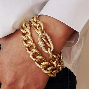 Könnte beinhalten: Goldfarbenes Armband-Set mit Kettengliedern. Ein Armband hat einen funkelnden, kristallbesetzten Verschluss. Die Armbänder sind dick und klobig, mit poliertem Finish. Der Schmuck wird an einem Handgelenk getragen, vor einem weißen Hemd und Jeans.