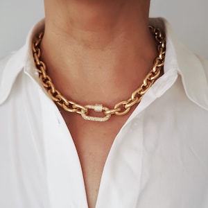 Op de afbeelding: Een goudkleurige ketting met een ontwerp van grote schakels. De ketting heeft een sprankelende, met diamanten bezette sluiting. De ketting wordt gedragen over een wit overhemd met knopen.