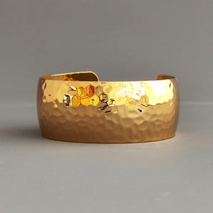 Puede incluir: Un brazalete ancho dorado con textura martillada. El brazalete tiene una forma ligeramente curvada y un acabado pulido, que refleja la luz. El brazalete mide aproximadamente 5 cm de ancho y está sobre un fondo gris.