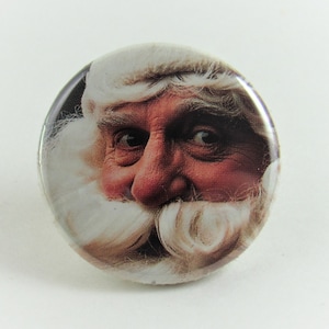 May include: Close-up of a round button featuring a Santa Claus face with a white beard and mustache. The button has a red background and a white border.