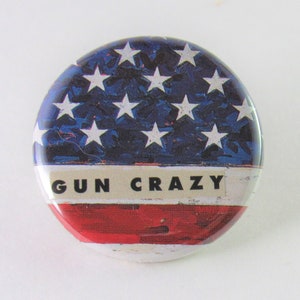May include: A round button with a red, white, and blue American flag design. The text "GUN CRAZY" is printed in black letters on a white stripe.